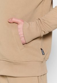 Beige sweatshirt med ribbestrikkede mansjetter og nederste kant. Har en sidelomme og en liten etikett på venstre side nær kanten. Myk tekstur.