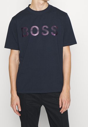 Mann trägt ein schwarzes T-Shirt mit kurzen Ärmeln mit glänzend violetten gestreiften Buchstaben, die "BOSS" über der Brust buchstabieren.