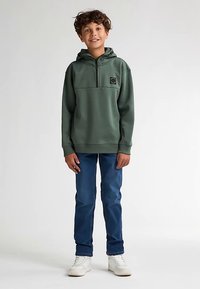 Zelená mikina s kapucí a polovičním zipem, velkou přední kapsou a černou nášivkou s logem, kombinovaná s modrými džínami a bílými teniskami.