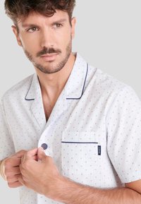 Camisa de pijama blanca con un fino patrón a rayas y puntos multicolores. Presenta cierre de botones, bolsillo y detalles de ribete azul.