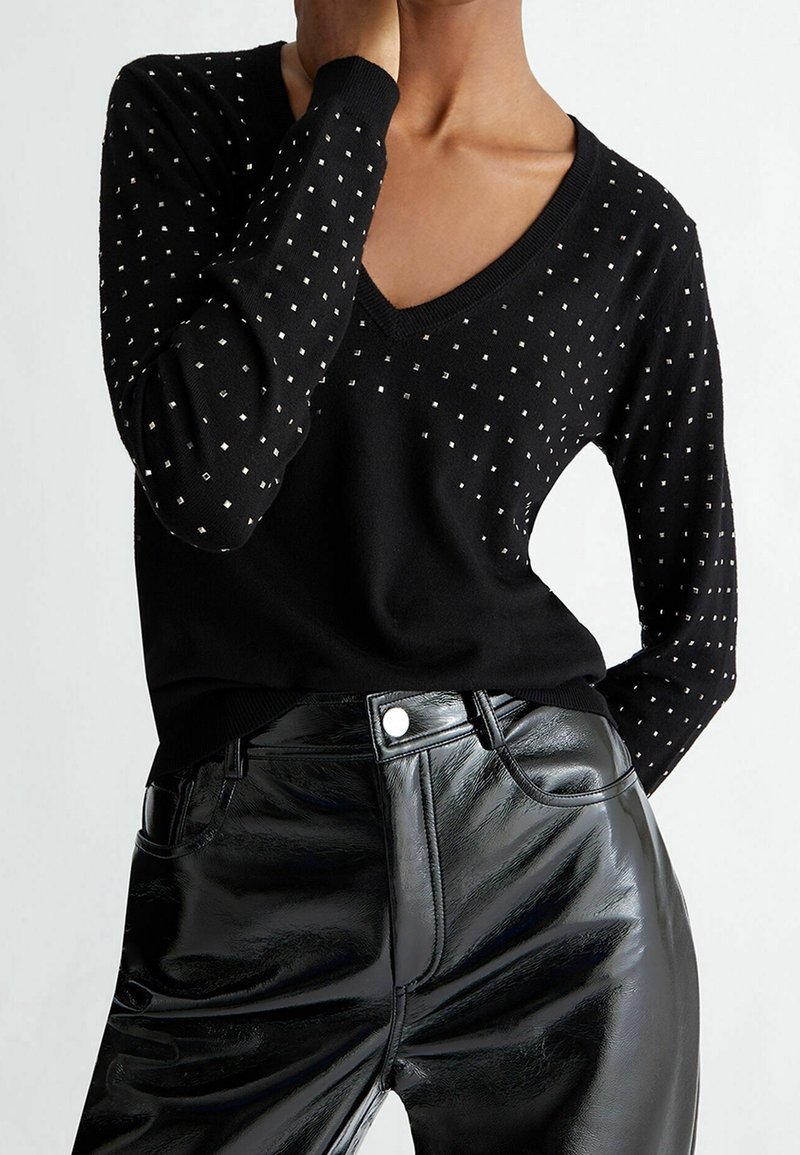 Personne portant un pull noir à col en V avec de petits motifs carrés blancs et un pantalon en cuir noir brillant taille haute, main près du cou.