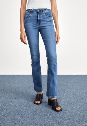 Kobieta w niebieskich dżinsach o średnim stanie i czarnych otwartych butach na obcasie, stojąca na niebieskim dywanie.