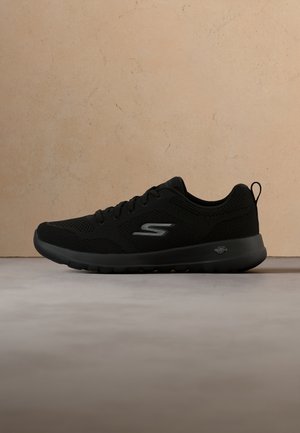 Czarny sportowy sneaker marki Skechers z siateczkową cholewką, sznurowaniem z przodu i amortyzowaną podeszwą, pokazany z profilu na szarej powierzchni.