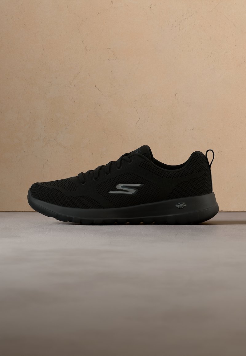 Baskets de sport noires Skechers avec dessus en mesh, laçage à l'avant et semelle rembourrée, vues en profil latéral sur une surface grise.