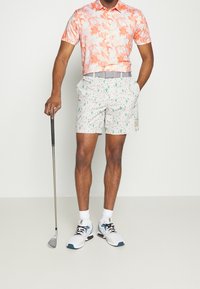 Polo s krátkým rukávem v oranžové květinové vzoru, bílé šortky se zeleným vzorem kaktusu, šedý pásek a bílé tenisky s tyrkysovými akcenty; drží golfovou hůl.