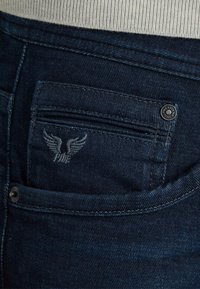 Tamno plave traperice sa stražnjim džepom s izvezenim logom orla, metalnim gumbom i šivenim detaljem na pojasu.