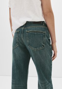 Temno teal jeans z visokim pasom, ki imajo zadnje žepke, subtilne madeže in kontrastne šive. Oblečeni z belo majico.