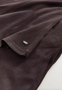 Tissu brun foncé doux montrant un gros plan d'une manche pliée avec une petite étiquette métallique rectangulaire attachée près du poignet.