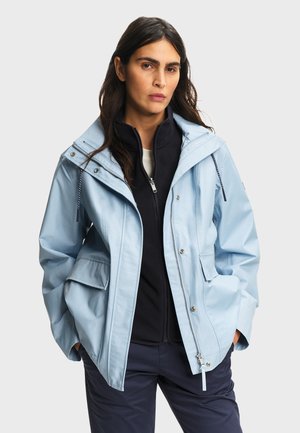 Femme aux longs cheveux bruns portant une veste bleu clair sur un polaire noir à fermeture éclair, debout les mains dans les poches de la veste sur un fond gris.