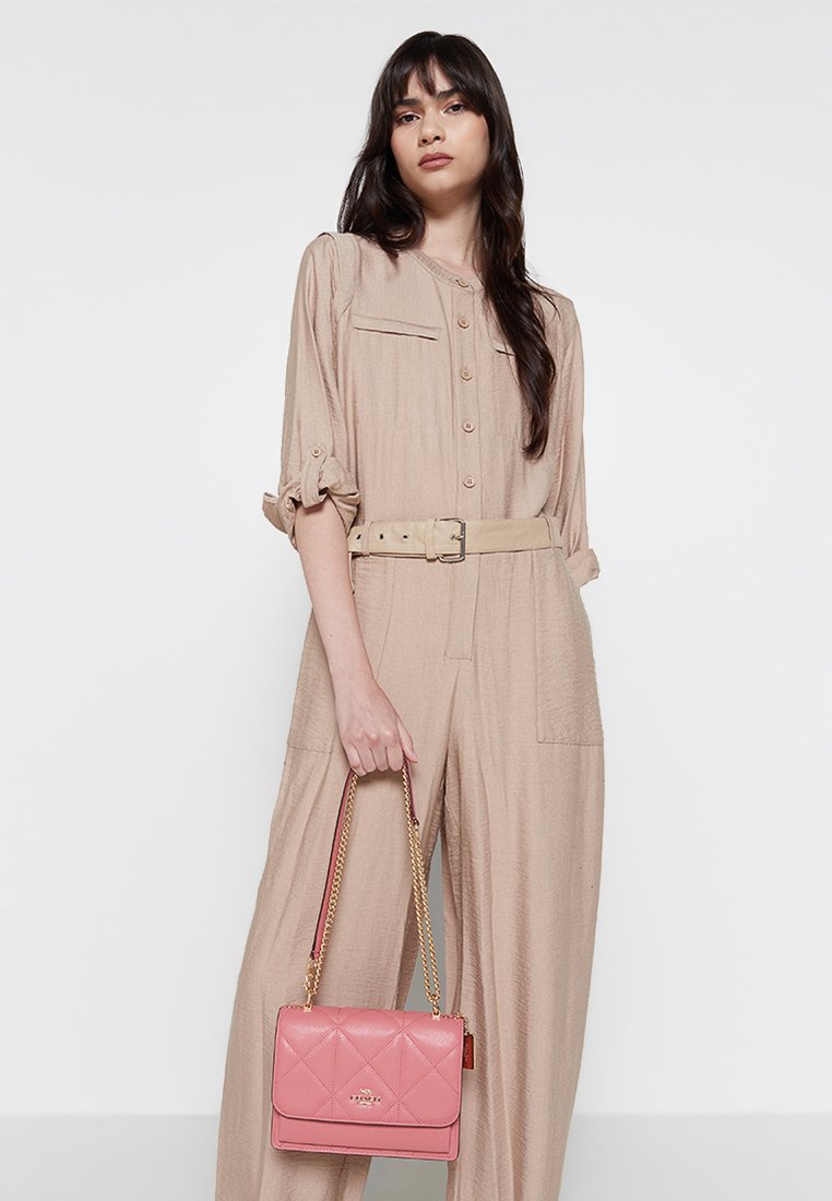 Mono beige con mangas estructuradas y cinturón, combinado con un bolso acolchado rosa que cuenta con una correa de cadena. Tejido suave y textura blanda.