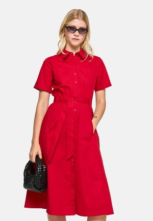Woman with long blonde hair wears black sunglasses, a bright red belted shirt dress, and holds a small black woven handbag.