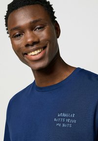 Tummansininen puuvillapaita, jossa on pyöreä kaula-aukko ja kevyesti sinisellä brodeerattu teksti: "WRANGLER BUTTS DRIVE ME NUTS." Sileä pinta.