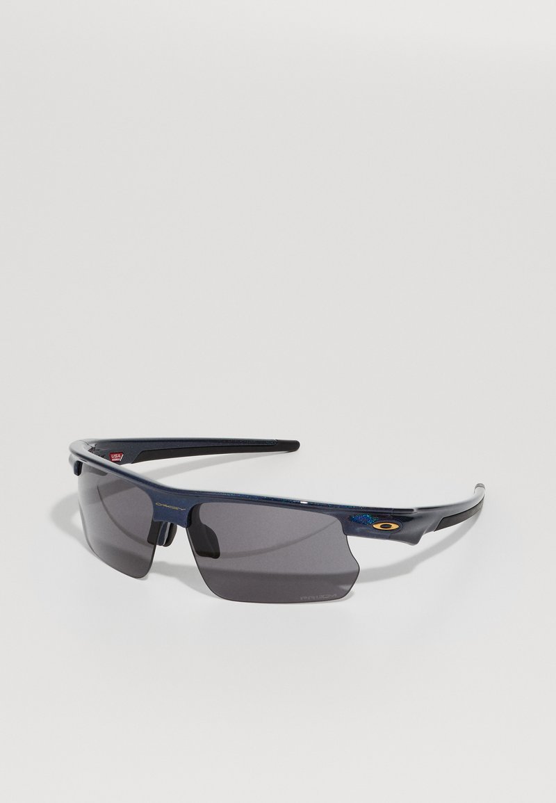 Gafas de sol con lentes gris oscuro, montura de plástico azul y un diseño aerodinámico y elegante. Presentan acentos con el logo y empuñaduras texturizadas en las patillas.