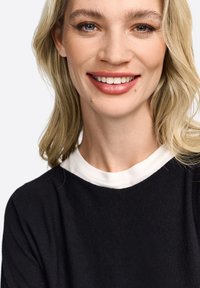 Close-up van een persoon met lichtblond haar, gekleed in een zwart top met een witte kraag. De top heeft een stevige textuur en een ontspannen fit.