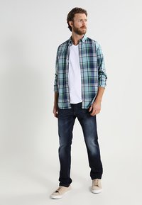 Karierter Button-up-Hemd in Grün, Blau und Weiß, über einem weißen T-Shirt; kombiniert mit dunklen Jeans und hellen Sneakern. Lässiger Schnitt, strukturierter Stoff.