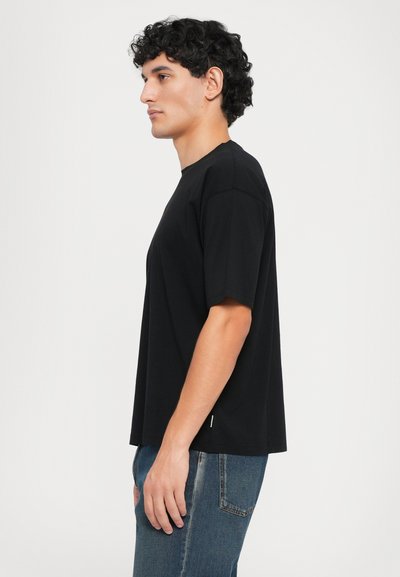 Svart bomull t-skjorte med korte ermer, rund hals og rett snitt. Kombinert med blå denimbukser, vist fra siden.