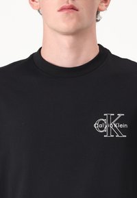 Μαύρη βαμβακερή μπλούζα με ριγέ λαιμόκοψη και λευκό κεντημένο λογότυπο της Calvin Klein στην αριστερή πλευρά του στήθους.