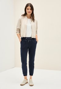 Veste bomber beige clair sur une chemise blanche, associée à un pantalon de jogging bleu marine avec poches et poignets élastiques. Des baskets décontractées complètent le look.