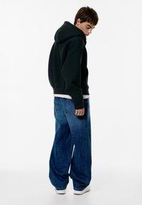 Felpa nera con cappuccio e vestibilità comoda, abbinata a jeans slim fit blu. Accenti di sneakers bianche, texture liscia.