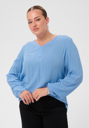 Femme aux cheveux plaqués en arrière porte une blouse bleu clair à manches longues bouffantes et un pantalon noir, accessoirisée avec des boucles d'oreilles en perles et des bracelets en argent.
