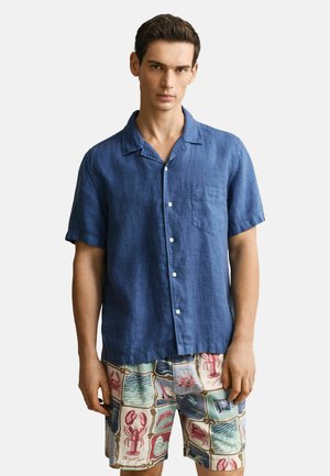 Man draagt een blauw overhemd met korte mouwen en kleurrijke korte broek met mariene en nautische ontwerpen, staand tegen een effen achtergrond.