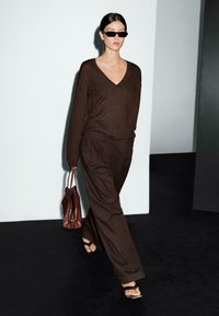 Bruine gebreide trui met V-hals, bruine geplooide broek, kleine donkere zonnebril en een bruine leren handtas. Model loopt tegen een grijze achtergrond.