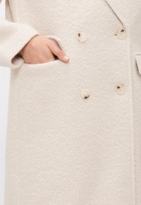 Beige ullblandingsfrakk med teksturert finish, som har doble knapper, store knapper og en sidelomme.