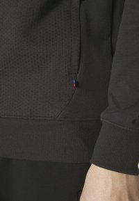 Sudadera negra con una tela texturizada, puños acanalados y una pequeña etiqueta tricolor en el bolsillo, con un diseño elegante y moderno.