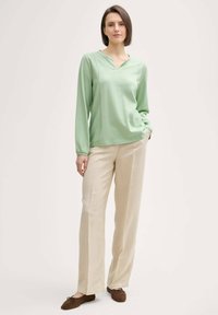 Haut à manches longues vert clair à côtes avec un col en V, associé à un pantalon ample beige. Des chaussures plates marron complètent la tenue.