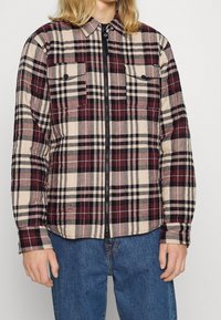 Camicia a quadri con tonalità di bordeaux, nero e beige. Dotata di zip sul davanti, due tasche sul petto e colletto. Tessuto morbido e strutturato.