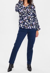 Blusa blu navy a fantasia con maniche a campana sopra pantaloni slim-fit blu scuro. Il modello indossa stivali neri con tacco, in piedi su uno sfondo neutro.