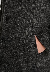 Gros plan sur un manteau en mélange de laine gris foncé, présentant un tissu texturé, deux boutons noirs et une poche latérale partiellement visible.
