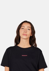 Černé bavlněné tričko s krátkými rukávy, na kterém je uprostřed malé červené logo "ROSSIGNOL". Jednoduchý a jasný design.