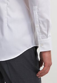 Biała koszula z długim rękawem z mankietami zapinanymi na guziki, wykonana z gładkiej tkaniny. Zaokrąglony dół i dopasowany krój widoczne obok ciemnych spodni w wzory.