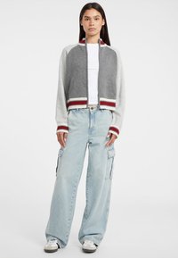 Giacca grigia e crema con maniche raglan, orlo a coste e tasche. Abbinata a pantaloni cargo a gamba larga di colore azzurro chiaro e scarpe da ginnastica bianche.