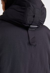 Schwarze Kapuzenjacke mit einer strukturierten, gepolsterten Kapuze und einem Metall-Schnallendetail auf der Rückseite. Glattes, leichtes Gewebe mit matter Oberfläche.