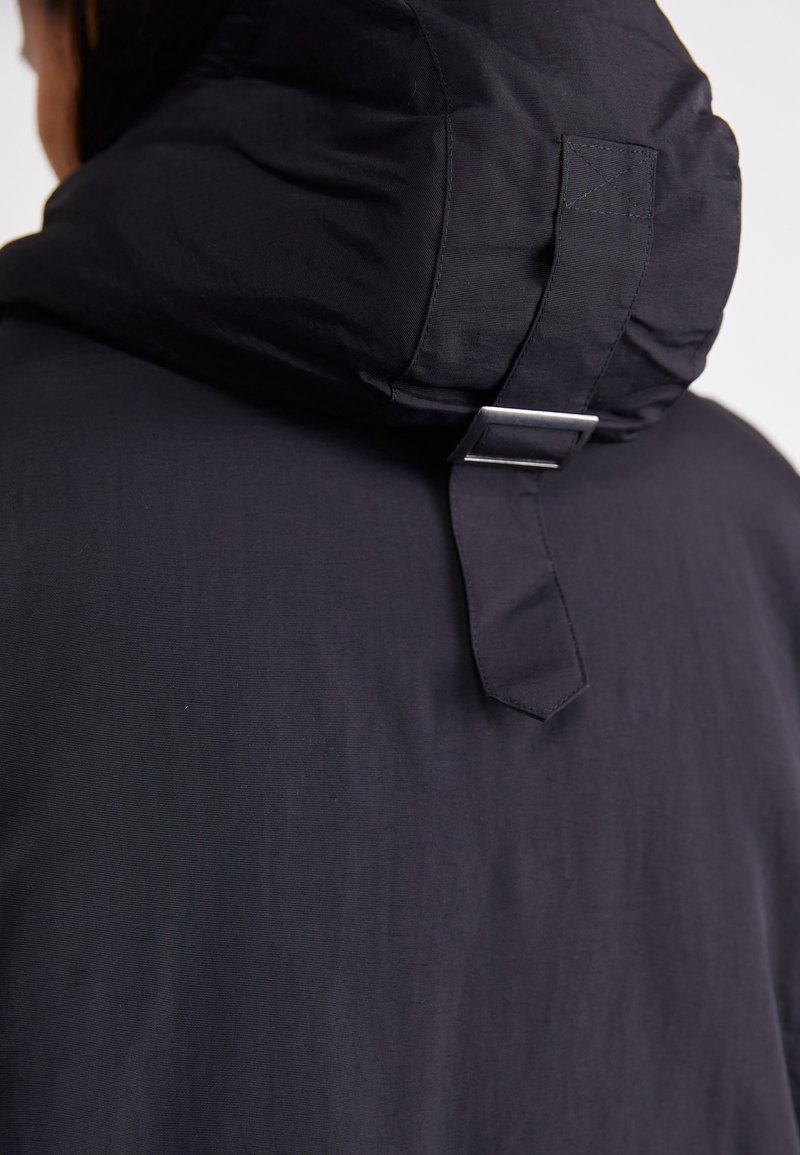 Schwarze Kapuzenjacke mit einer strukturierten, gepolsterten Kapuze und einem Metall-Schnallendetail auf der Rückseite. Glattes, leichtes Gewebe mit matter Oberfläche.