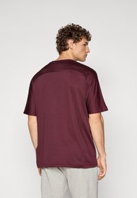 Bordeaux kortarmig T-shirt van gladde stof, met een ronde hals en een losse pasvorm, gedragen met grijze joggingbroek.