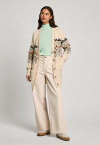 Cardigan crema con dettagli a motivo in arancione e blu, indossato sopra un top a costine verde menta e abbinato a pantaloni larghi beige chiaro.