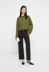 Túlméretezett olajzöld pulóver, fekete, bőszárú farmernadrág, fekete, hegyesorú magassarkú cipő, kezében egy kis fehér kézitáska. Minimalista dizájn és semleges színpaletta.