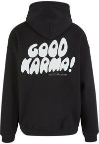 Fekete kapucnis nadrág, amely pamutból készült, és a hátsó részén egy nagy fehér buborékbetűs felirattal rendelkezik: "JÓ KARMA!"