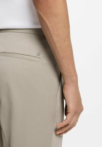 Beżowe spodnie z gładkiego materiału, posiadające tylną kieszeń i mały detal z logo marki. Ręka spoczywa blisko biodra, widoczna minimalna tekstura.