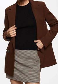 Blazer marron sur mesure porté sur un haut noir côtelé, associé à une jupe courte à motif pied-de-poule. Il présente des revers pointus et deux boutons à l'avant.