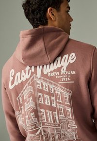 Bruklino spalvos kapučino su baltu grafinio dizaino pastatu, kuriame užrašyta "East Village Brew House", su virvelėmis reguliuojamu gobtuvu ir minkšto paviršiaus.