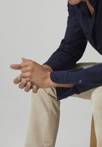 Persoon in een marineblauwe shirt en beige broek, zittend met de handen clasped en rustend op één knie tegen een effen achtergrond.