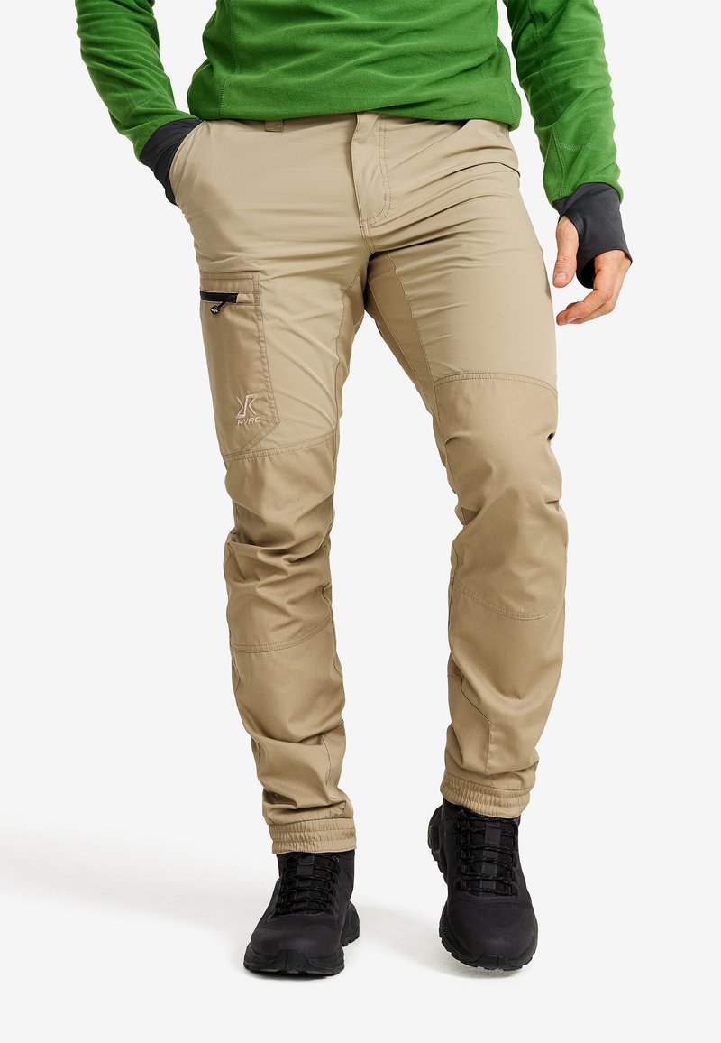 Beige outdoor pants with zippered side pocket, elastic cuffs, and reinforced knees, paired with black hiking boots and a green top.
