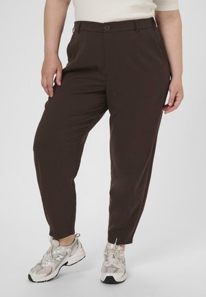 Person wearing dark brown tapered pants and white athletic shoes, standing on a plain light background.