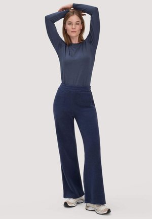 Woman wearing navy long-sleeve top and navy wide-leg pants stands with arms raised above head, wearing white sneakers.