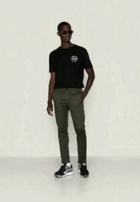 T-shirt de algodão preto com gráfico branco, calças slim-fit verde-oliva, e ténis pretos com detalhes brancos e cinzas, acompanhados de óculos de sol.