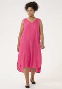 Vestido sin mangas de lino rosa con escote en V, dobladillo asimétrico y corte relajado, combinado con slippers negras sobre un fondo blanco liso.